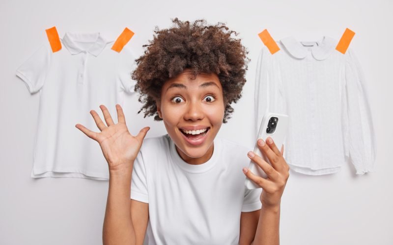 Excited emotional young woman with cheerful expression keeps palm raised reacts happily on amazing news uses mobile phone dressed casually isolated over white background with clothes behind.